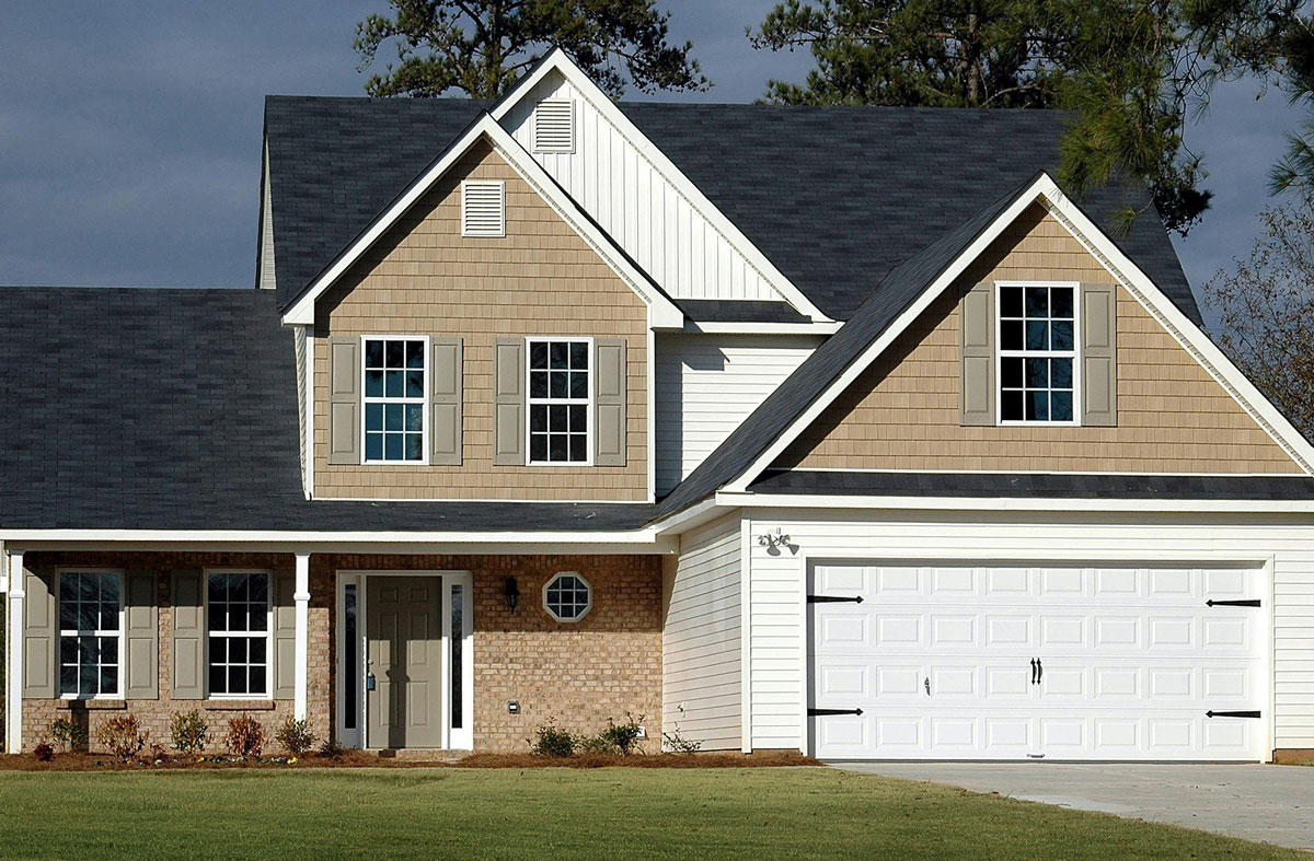 A two-story house with a beige exterior and white trim. The house features a covered porch and a two-car garage.