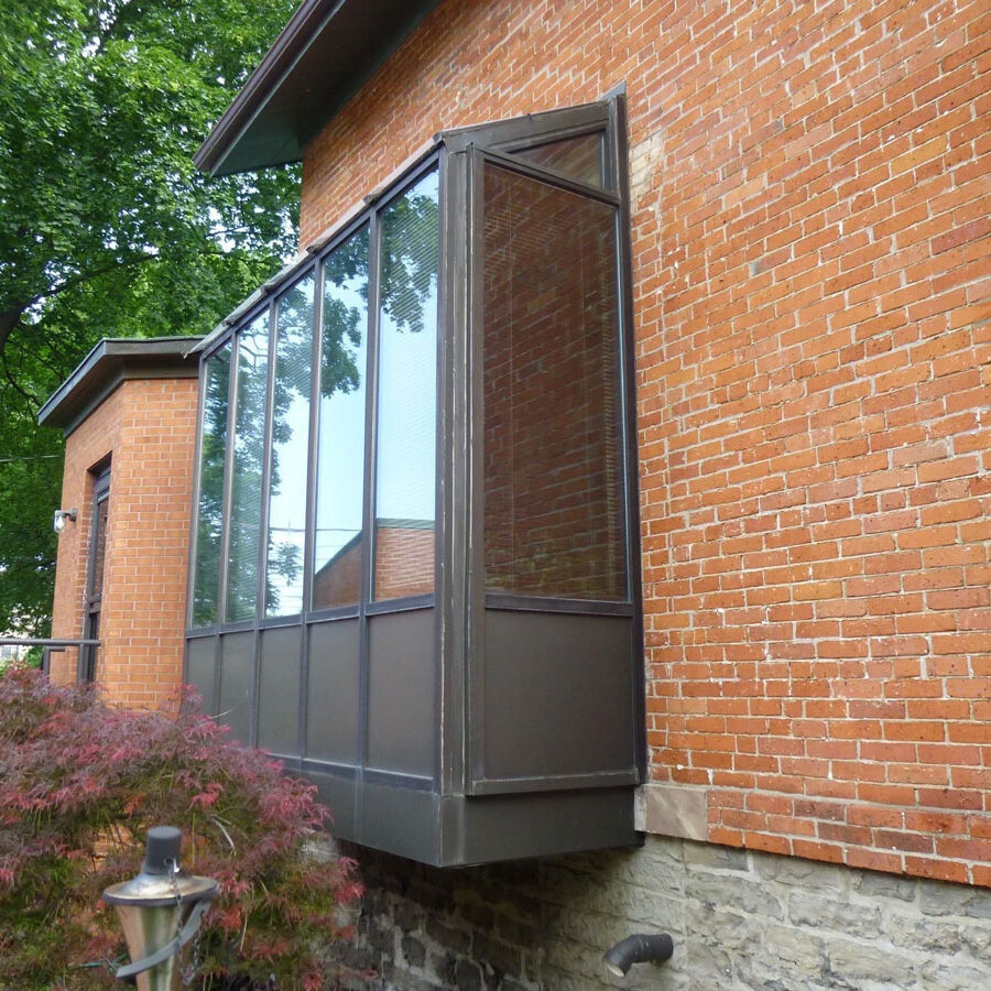 A sunroom with glass panels on a red brick house. The sunroom has a brown frame and is attached to the side of the house.
